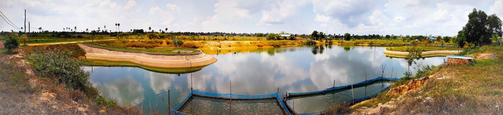 Photo shows an artificial lake with small fishponds in flexible fishnets. The landscape is the redyellowbrown of the rich soil of Cambodian countryside. The lake mirrors a cloudy sky with some clear blue spots.
