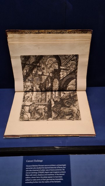Display cabinet, glass fronted, large ancient tome spread open showing a Giovanni Piranesi Carceri etching, a strange prison fantasy landscape, incredibly detailed, black and white. 