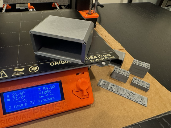A 3D printed box made from separate printed panels. It sits on a 3D printer, and previous test prints of the Prusa logo and LEGO bricks are next to it. 