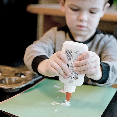 A young boy attempting a two-handed squeeze to apply glue from a squeeze bottle onto a sheet of paper.