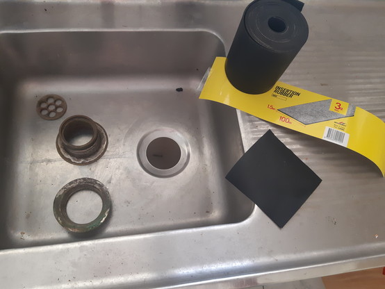 Old stainless steel sink with roll of  nitrile rubber sheeting and sink waste fittings sitting in/on it. A square of the rubber sheet has been cut from the roll. It will be used to make the seal.