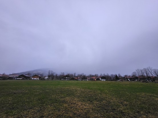 This image presents a serene, expansive grassy field bathed in natural light, extending towards a picturesque background adorned with houses, subtly hinting at the interface between rural charm and human habitation. The sky, a canvas of clouds, ranges from fluffy formations to denser clusters, suggesting a day of variable weather, potentially in a season such as late winter or early spring given the lushness of the grass and the bareness of some trees. The landscape is a harmonious blend of nat…