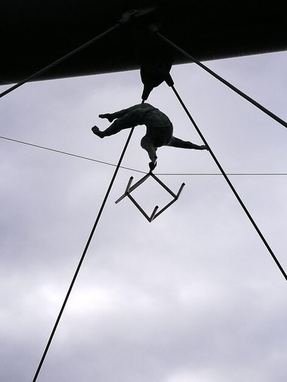 Stunt person up in the air bending backwards holding onto wire with one hand and holding a chair in the other hand