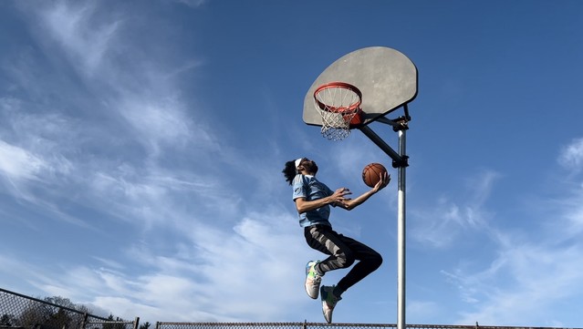 Guy in a light blue top and black sweatpants with an urban camouflage stripe down the side goes up for a left-handed reverse layup from the left side of the hoop to the right. The sky is blue with scattered, wispy clouds. 