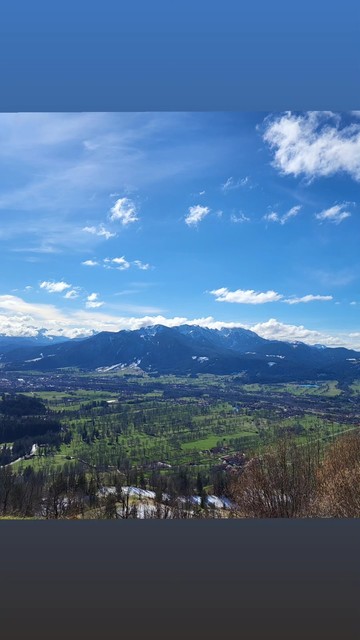 The image depicts a serene valley scene, transitioning between seasons. Bare trees frame a tapestry of green fields and dark woods, punctuated by distant buildings. Snow-capped mountains rise in the background, shrouded by clouds, while a vibrant blue sky with fluffy clouds completes this tranquil vista. The landscape exudes peace and natural splendor, capturing the essence of a picturesque, idyllic setting.
