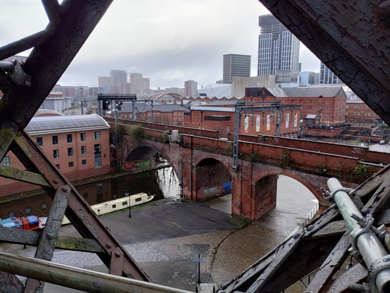A view through iron girders of a brick viaduct with several buildings, and a canal with a narrow boat.  Some high rise buildings in the distance.