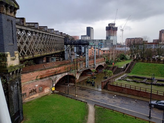 An iron girder viaduct crossing over a brick viaduct with a road going under both. A grassy area on the right with ruins of the old Roman fort. Some high rise buildings in the distance.