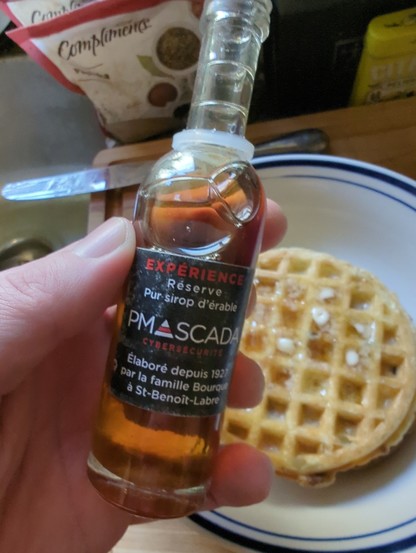 It's a sample sized bottle of maple syrup from the "famille Bourque a St-Benoit-Labre" branded with the logo of PM Scada Cybersecurite with Eggos in a bowl behind