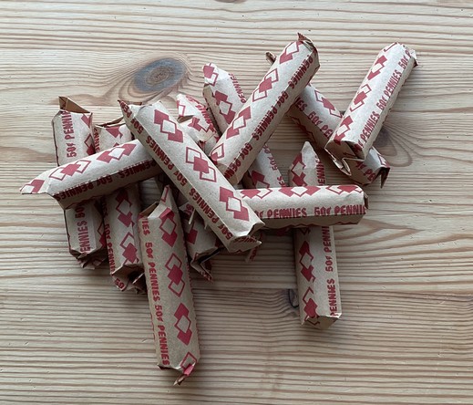 Thirteen rolls of pennies on a wooden table top. The pennies are wrapped in kraft wrappers printed in red.