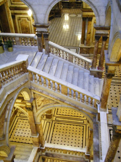 Looking down on a complicated free-standing staircase made of white marble with a thick baroque balustrade, many arches and painted columns. The overall tone is golden. 