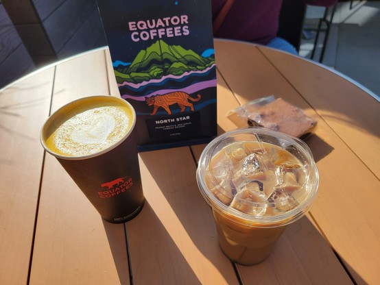 Outdoor cafe table with light-brown medium-width slats. On the table is a coffee pouch of Equator Coffees North Star coffee beans, a 16-ounce paper to-go cup with a coffee turmeric latte with a heart in the milk foam, a brownie in plastic wrapper, and an iced coffee latte.