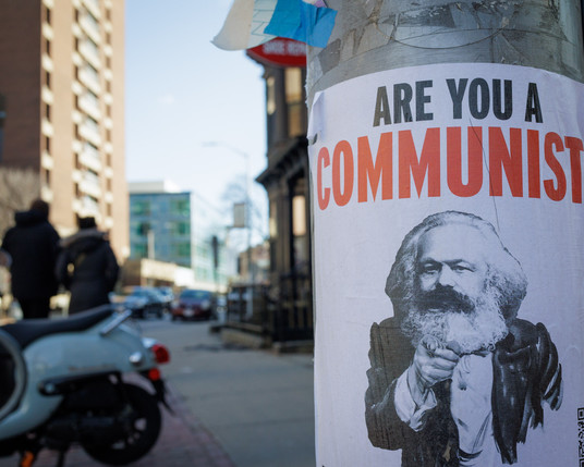 Close up color photo of a poster glued to a utility pole. The visible text of the poster reads “ARE YOU A COMMUNIST” with the image of Karl Marx below posed with his finger pointing at the viewer in the same way an Uncle Sam poster would with the saying “I WANT YOU.” To the left, and out of focus is a street scene with the back half of a parked scooter, two silhouetted figures, and  some apartment buildings.