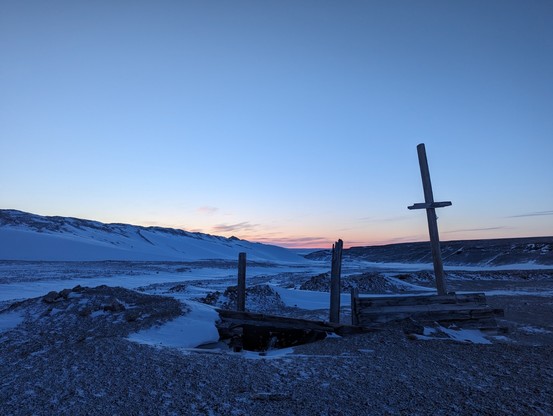 Three wooden poles stick up above an old wooden entrance to a bunker. The ground is made up of glacial moraine material. There is snow everywhere. A large mountain is off to the left behind the wooden poles and it has huge walls of snow. The sky is in twilight even though it is the middle of the day.