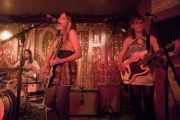 Singer/guitarist and bassist on stage with a drummer in the background in front of the gold ribbon backdrop of the MOTH Club. The singer, Sadie Dupuis, has her mouth wide open as if she is singing "ahhhh" or similar.