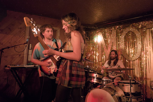 Singer/guitarist on stage facing other guitarist with drummer in the background.
