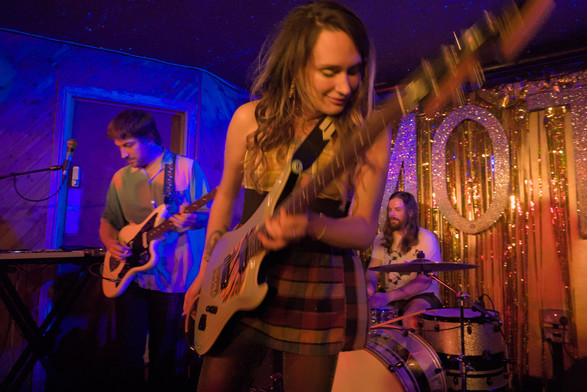 Slightly blurred, due to slow shutter and motion, guitarist with next of guitar towards camera. Another guitarist and drummer in background. 