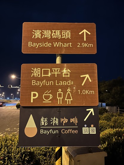 At night looking at some new signs arranged one atop the other on a striped yellow and black pole off the right side footpath where some bushes are.  The top two signs are brown with direction arrows pointing up and slightly right, while the bottom sign is dark grey with an arrow pointing further right, and all have light coloured lettering.  The top sign has a typo of “Whart” instead of “Wharf”.  They read:

濱灣碼頭 Bayside Whart 2.9Km
潮口平台 Bayfun Land 1.0Km (with parking, coffee and toilet icons underneath)
戲湖咖啡 Bayfun Coffee (with a water drop shaped logo on the left and Facebook and Instagram QR codes on the right)