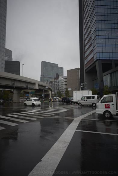 A busy traffic intersection in the day. The road is slick with rain. A raised freeway curves in from the left to the centre of frame, then back out into the distance. At the peak of the curve, in the distance, is Nakagin Capsule Tower - a stack of cubes, with round windows, dwarfed by surrounding buildings.