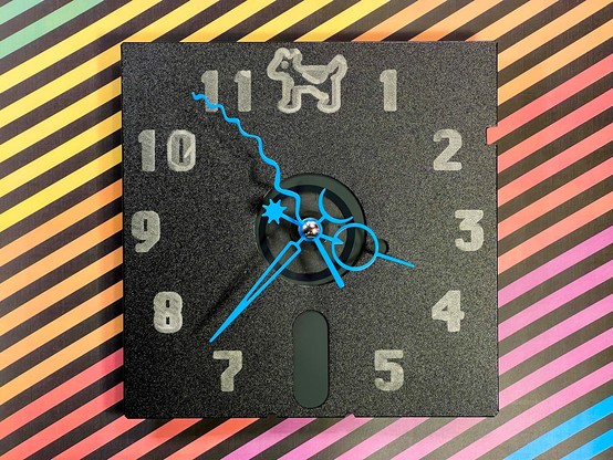 A 5.25” black floppy disk with blue clock movement arms in the center with numerals pen plotted in silver in Chicago font with smooth Clarus at the 12 position. It’s on a background of Apple logo color stripes. 