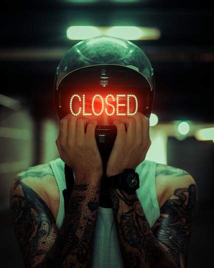 Tattooed man wearing motorcycle helmet with "CLOSED" in red digital letters where the face mask should be