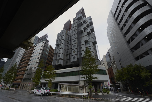 A photo from under an overhead expressway, looking up at Nakagin Capsule Tower. The building is a geometric collection of cubic blocks, in a semi-organic arrangement, each block has a round window, half the diameter of the entire face. There are two pointed spires, one higher than the other, atop the two main masses of the building, and a large plinth base, floating a story above street level. The plinth has a long window running along its length, as if it’s a single big enclosed communal space for the residents.

On one window, someone has stuck up pies of paper, triangles for eyes, and a row of teeth, making the window look like a giant jack-o-lantern, as Halloween was in two days.