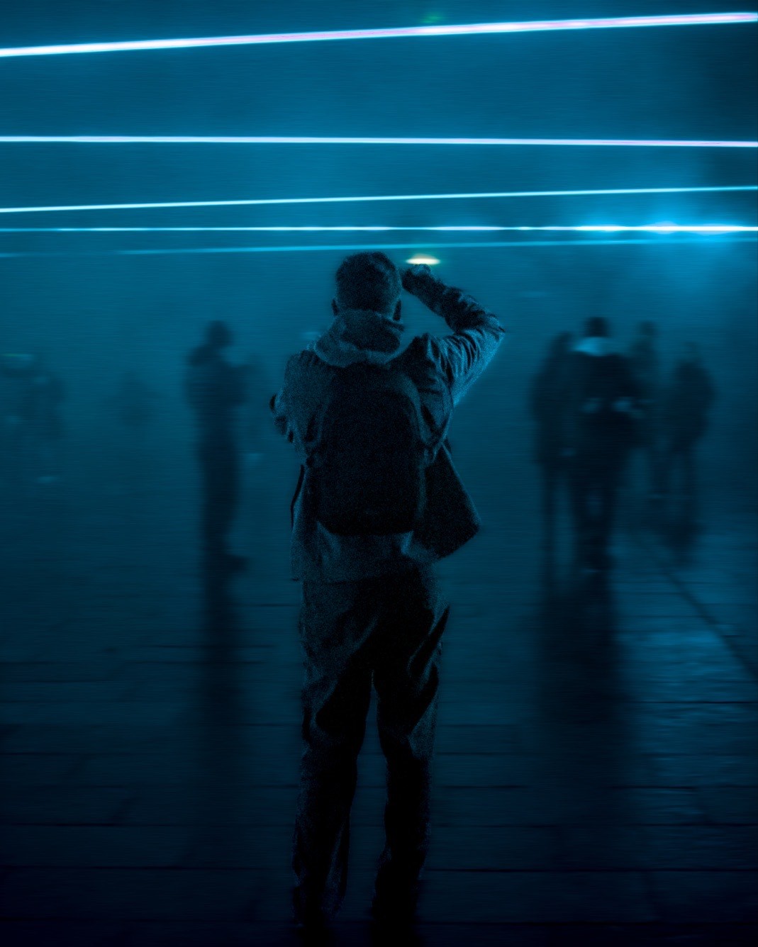 This image features a person seen from the back, standing in a dimly lit space illuminated by horizontal neon blue lights. The person is holding what appears to be a camera, taking a photo. Shadows and silhouettes of other individuals are visible in the background, suggesting a public or social setting.