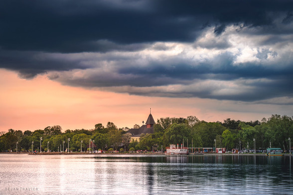 A view of the beautiful Palic lake in Serbia one spring day while the clouds slowly hover over it. There are still little nice warm tones left in one part of the sky. In the distance, you can see The Great park Palic with the Grand terrace in the middle surrounded by trees. we also see a promenade by the lake with a lot of people walking as well as some of the sights of the Palic nature park.

Palic lake is the perfect place for everyone looking for a pleasant holiday on the lake, walks in nature and good entertainment. This is one of the most visited destinations in Serbia, primarily due to its beauty and diverse possibilities for holiday and relaxation. legend says that the lake is a remnant of the Pannonian Sea, that existed in this region in ancient times. It is a lake that has been one of the most visited locations in Serbia for many years, primarily because of its landscaping, beauty and various opportunities for rest and recreation. Palic Lake is located in Vojvodina, 8 kilometers away from Subotica, one of the most beautiful cities in Serbia, so all visitors who visit the lake can easily spend their time in this city as well. It is one of the most beautiful lakes in Serbia and the largest natural lake in Serbia as well.
One May day on Palic lake, Vojvodina, Serbia. From the Lakes collection.
