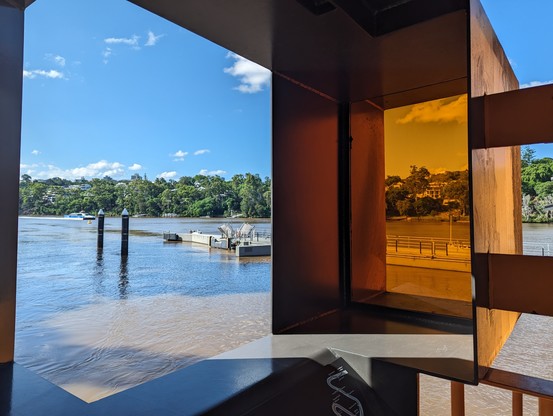 Brisbane River, looking South from the St Lucia ferry terminal.
Scene is divided by a bronze-tinted window to the centre. To the left, in the distance under the blue sky reflected in the water, a ferry is arriving.