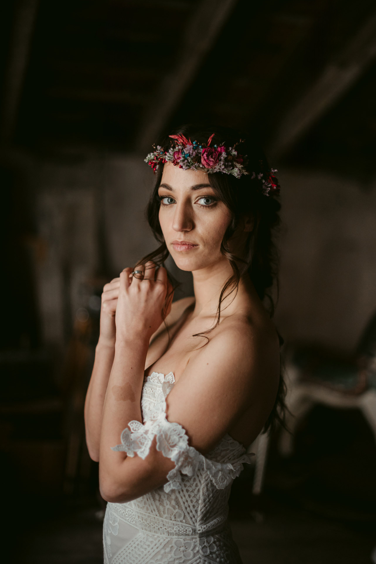 The image features a bride in an elegant, understated lace wedding dress, standing in a room with subdued lighting that creates an intimate and contemplative atmosphere. Her gaze appears to be directed into the distance, exuding calmness and confidence. She wears a striking, colorful floral crown on her head, which forms a beautiful contrast to her dark hair. The natural light accentuates the texture of the lace dress and the soft details on the sleeves. The bride's pose is relaxed, with her hand gently touching her neck, conveying a sense of vulnerability and grace. The background is blurred and unobtrusive, drawing focus to the bride and her delicate features.
