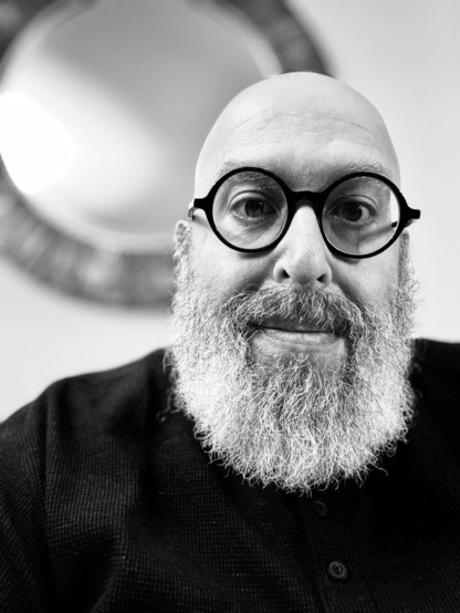 A picture of a bald, bearded man in black and white with round glasses and a mirror behind him
