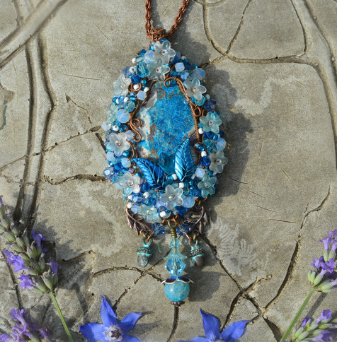 A light blue and white pendant necklace, teardrop-shaped. The central stone is sky blue mottled with grey and white. Surrounded by beadwork and curls of copper wire. Leaf and flower beads dot the pendant, acorns and a blue glass pendant hang underneath.