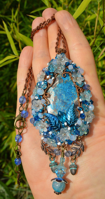 A blue and white wire-wrapped pendant. The central stone is blue, with an elaborate bread and wire surrounding, sewn through with tiny glass beads, flowers and crystals. The pendant is displayed on a hand, with greenery behind.