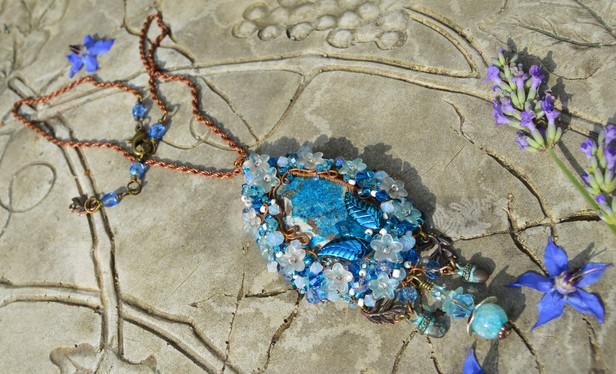 Sky blue pendant necklace, encrusted with beads, crystals and tiny flowers. The pendant and long chain are lying on a weathered stone table, with lavender and borage flowers around it.