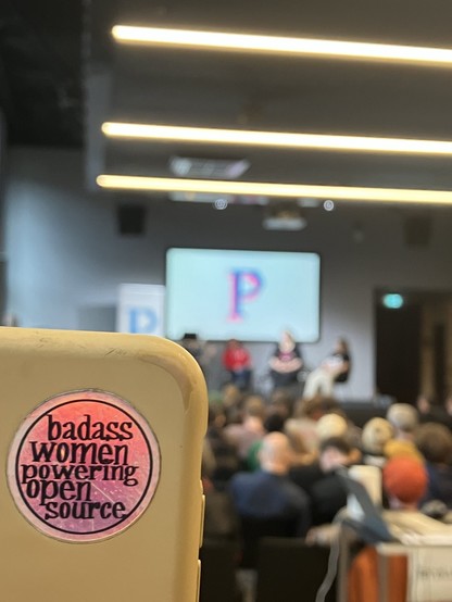 Sticker: "badass women powering open source", im Hintergrund eine unscharfe Bühne