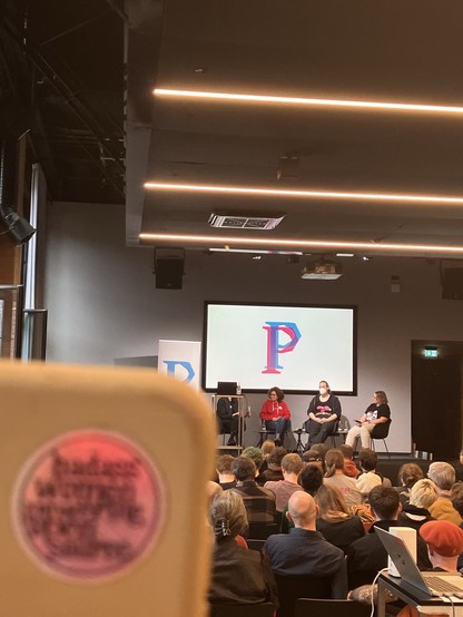 Ein unscharfer Sticker im Vordergrund, im Hintergrund eine Bühne mit Karo, Nadine und Marta.