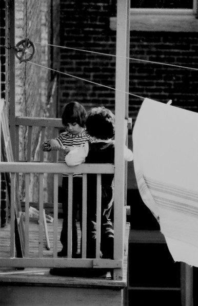 Black and white photo of two small kids playing on a wooden balcony, part of a brick building. There is an old fashioned pulley and cord laundry line, with a Hudson Bay blanket hung on it to air.