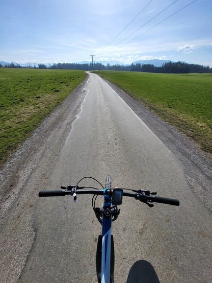 This image captures a serene outdoor scene featuring a bicycle positioned on a road. The road is surrounded by an expanse of grass, contributing to the peaceful ambiance of the setting. The sky, visible in the backdrop, adds to the picturesque quality of the scene with its clear and vast expanse, suggesting a fine day. The bicycle, the central object in the frame, is detailed and appears to be parked, hinting at a momentary pause in travel. The presence of wheels and the overall structure of th…
