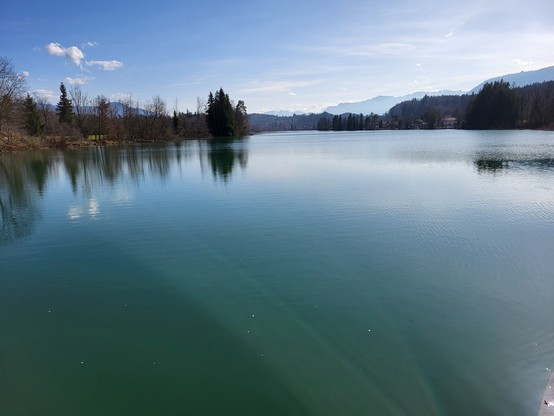 This image captures a serene and picturesque landscape that showcases the tranquil beauty of nature. In the foreground, the calm body of water acts as a perfect mirror, reflecting the lush trees that line its banks, creating a symphony of greens that enhance the scene's tranquility. Beyond the trees, majestic mountains rise into the sky, their silhouettes adding a sense of grandeur and scale to the composition. The sky above is adorned with soft, wispy clouds, suggesting a peaceful day. The ove…