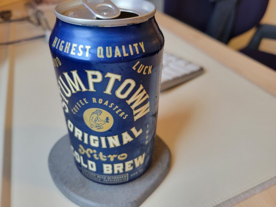 Can of Stumptown Original Nitro Cold Brew Coffee on a gray diatomaceous-earth coaster on an off-white desk mat on a maple desk. 

Can art description:
The overall effect is a maximalist, oldy-timey advertisement.
Solid metallic-blue background. The can has a 1/2-inch shoulder at top. Text on the shoulder reads "Highest Quality" in yellow block letters.
Below this on the main can face:
In the top corners, words "Good" and "Luck". Text "Stumptown" appears in a half-circle at top. Inside it in a smaller half-circle is "Coffee Roasters". Beneath this is an illustration of a naked person kneeling and holding a branch (presumably coffee berries) in one hand and a horseshoe in the other hand. Below this is text "Original", then "Nitro Cold Brew" in large letters. To the left and right is text "Estd. 1999". 
Footer text: 10.3 Fl Oz, "Infused with Nitrogen / Perishable Refrigerate", and 305 mL.