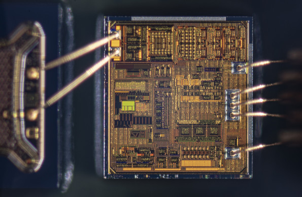 A bare IC with golden bond wires going to a blurry structure to the left that sits closer to the camera and another bundle of bond wires going down on the right of the IC.
The IC has very delicate structures and patterns and shimmers in a golden bronze hue in most places with some small blotches of green and red in some small areas.
