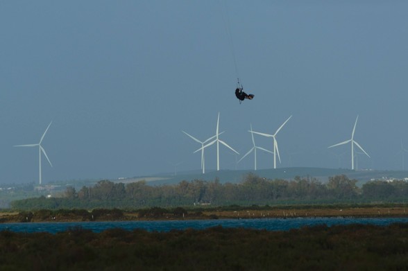 Un surfista que parece estar "volando" con su kitesurf sobre el caño de Sancti Petri, por encima de los generadores de energía heólica que se ven al fondo de la imagen.
