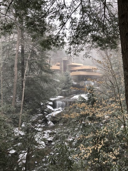 Winter photograph of Frank Lloyd Wright’s Falling Water from downstream.

The river and ground are partly covered with snow.
The house is visible through a gap in the trees; the colour of the leaves of one of the trees in the foreground matches that of the building.
