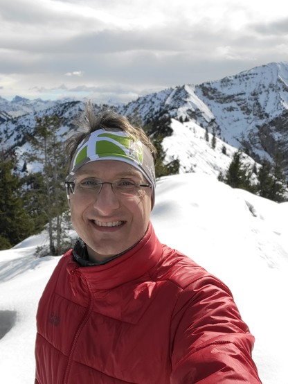 In the image, a man is captured in the midst of taking a selfie against a breathtaking backdrop of a snowy mountain landscape. The scene is predominantly swathed in shades of white and grey, reflecting the wintry setting. The man is dressed appropriately for the cold, outdoor environment, indicating a hiking activity. His attire includes goggles, suggesting preparation for skiing or protection against the glare of the snow. He is smiling, adding a warm, human touch to the chilly, majestic setti…
