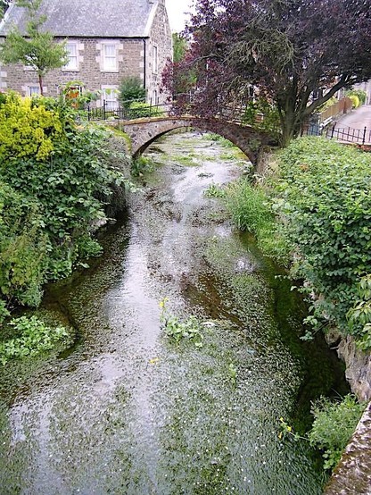Colour photo of a brook. There is an old stone bridge crossing it, abundant greenery and an typically Scottish stone house.