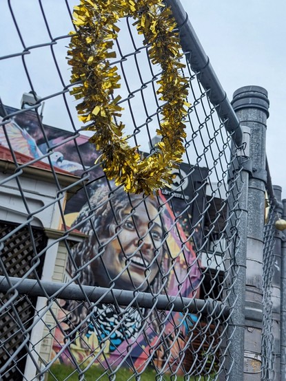 Shot is taken through a high mesh fence, which has a circle of golden Christmas tinsel affixed.
We see a brown skinned lady smiling. She is wearing a Palestinian keffiyeh around her neck and a colourful scarf with the Aboriginal flag's black yellow and red prominent.
