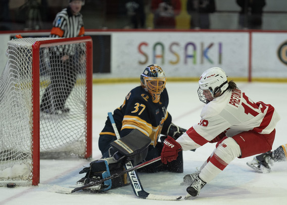 A female hockey goalie in a dark uniform with yellow trim and lettering reading "37"  physically blocks the goal behind her,  looking carefully in the lower left corner note the puck has passed by the edge of the goal.  The attacker is in a red and white clothes with the number "19".  Behind the goal note the referee with a striped shirt,  an ad for something called SASAKI, and I think a video crew on the other side of the glass on the far side.