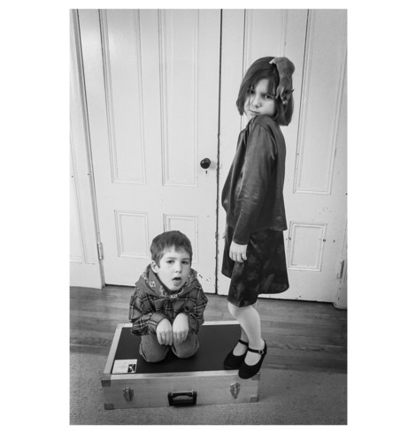 Black and white portrait of two children. One, a boy, is kneeling down on an equipment case pretending to be some kind of small animal with his hands pulled in close to his chest. He also has his mouth open with his tongue partially out. The other, a girl in a blouse and skirt is standing on the other side of the case turned a bit to the side, her face tucked down to her shoulder. She has a dark grey pet rat clinging to the side of her head with its head pointed down. The background is an old painted double-door that is closed behind them. 