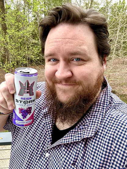 Me, a man with short hair and a beard. I'm wearing a button-up shirt that has a checkerboard pattern on it, and holding a can of Reign Storm Harvest Grape energy drink.