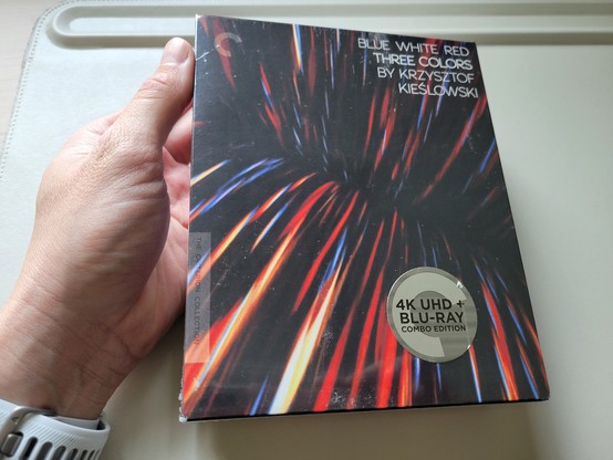Photo of the front of an unopened box set of The Three Colours trilogy (French: Trois couleurs, Polish: Trzy kolory) from The Criterion Collection, held in a hand over an off-white desk mat. The cover art shows the interior of a mass of red and blue wires, separated into an upper and lower half. 
