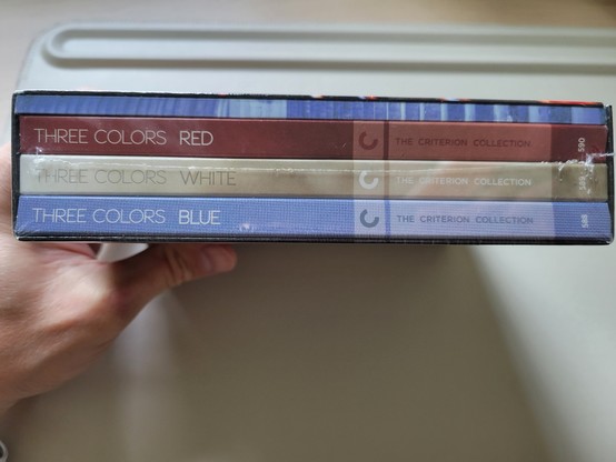 Photo of the spine of an unopened box set of The Three Colours trilogy (French: Trois couleurs, Polish: Trzy kolory), held in a hand over an off-white desk mat. The titles of the 3 movies are visible (from bottom to top): 1) Three Colors: Blue; 2) Three Colors: White; and 3) Three Colors: Red. The effect is to represent the colors of the Flag of France.